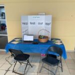 A photograph of the booth set-up. There's a table and two chairs, a basket full of snacks, a laptop computer, and a sign that says "Help test our app!"