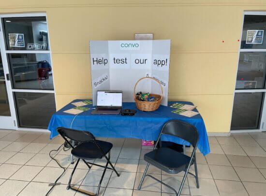 A photograph of the booth set-up. There's a table and two chairs, a basket full of snacks, a laptop computer, and a sign that says 