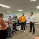 A photograph of the product squad in a standup meeting. Several team members are standing in a circle in the middle of an office.